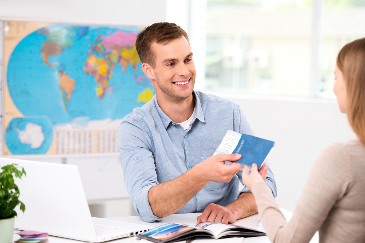 Photo of male travel agent and young woman. Young man smiling and giving tickets, passport with visa to female tourist. Travel agency office interior with big world map